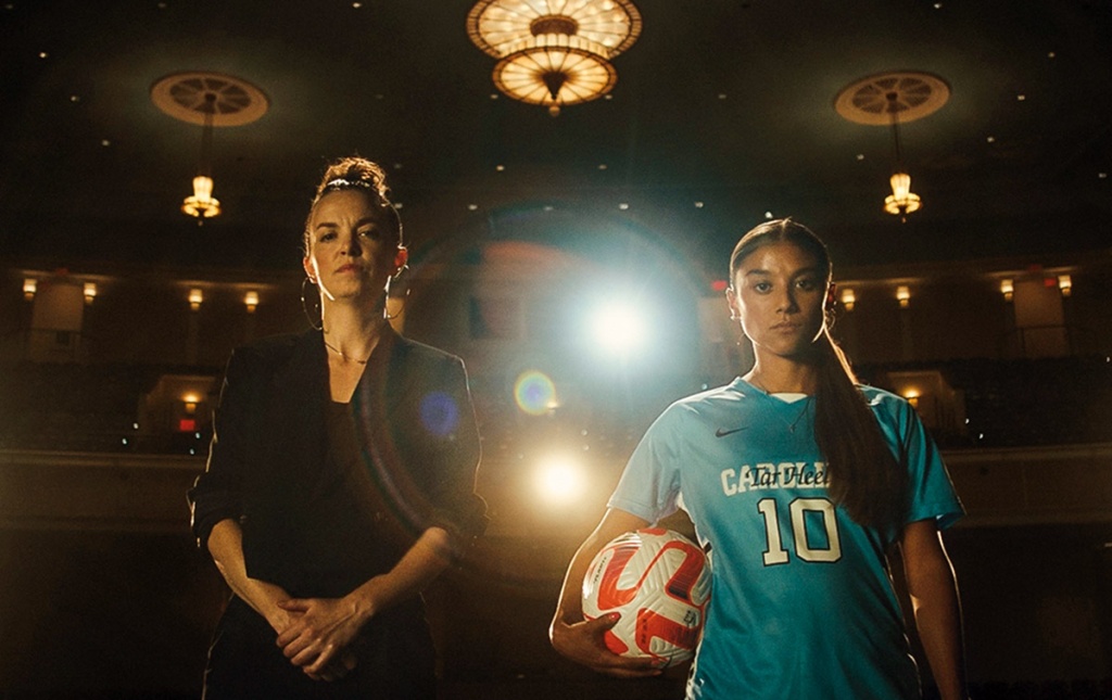 Image of two women standing on a stage with spotlights behind them, one holding a soccer ball.