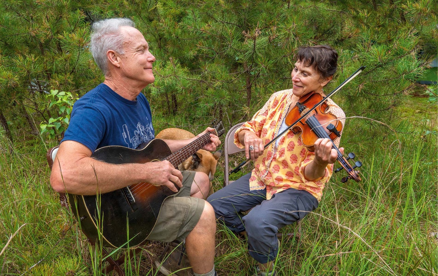 Rory MacLeod and Sandol Astrausky playing the guitar and fiddle in a green field by trees.