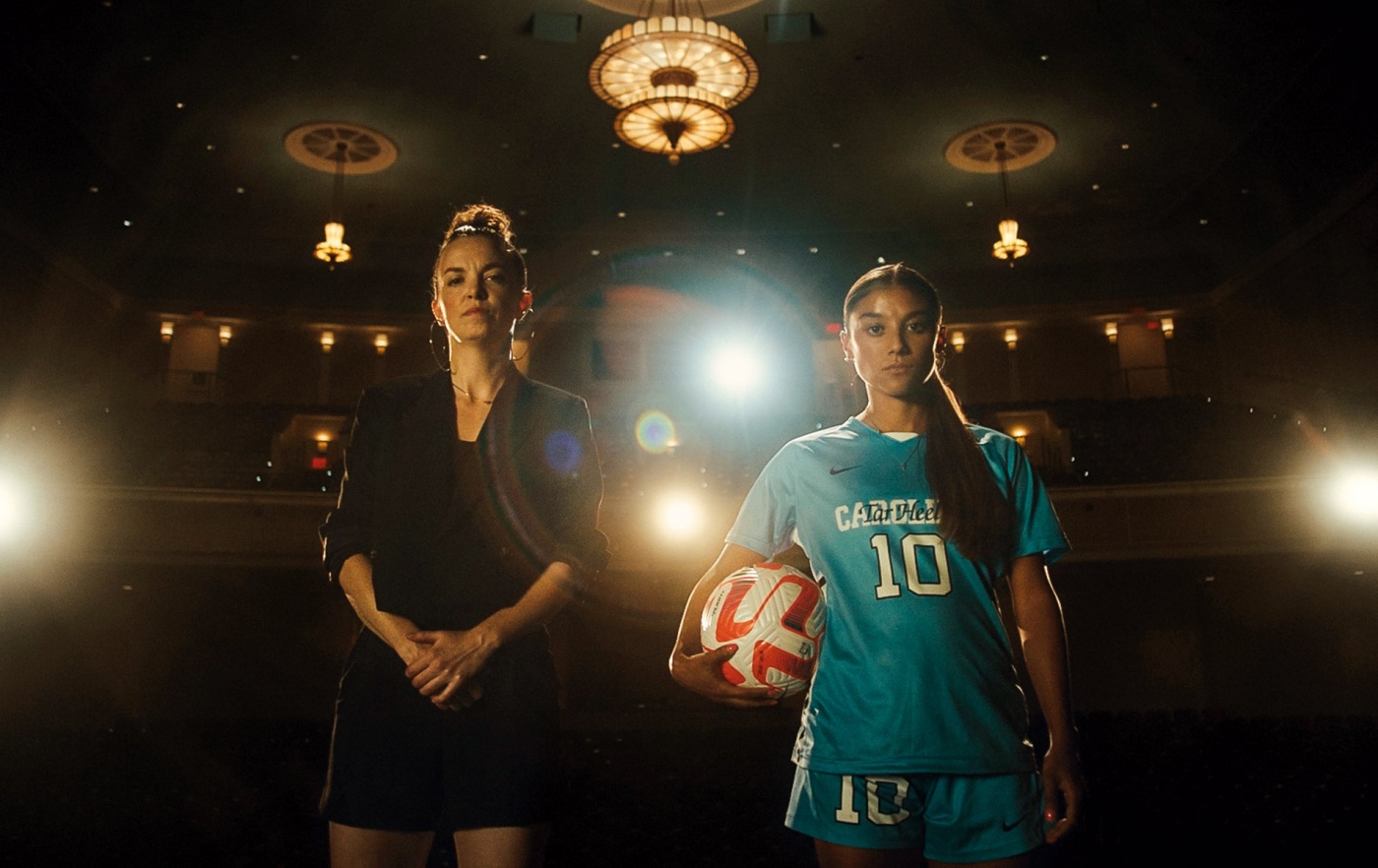 Image of two women standing on a stage with spotlights behind them, one holding a soccer ball.